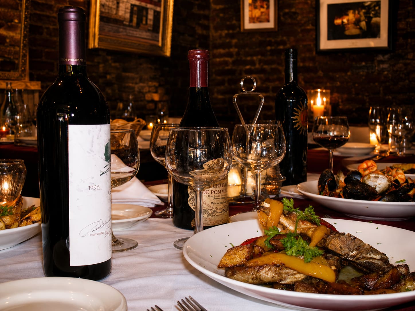A table mid-meal against an exposed brick wall hung with gilt-framed pictures — candles lit, several bottles of red wine open, glasses poured, a plate of chop with peppers and potatoes and a bowl of shellfish still going.