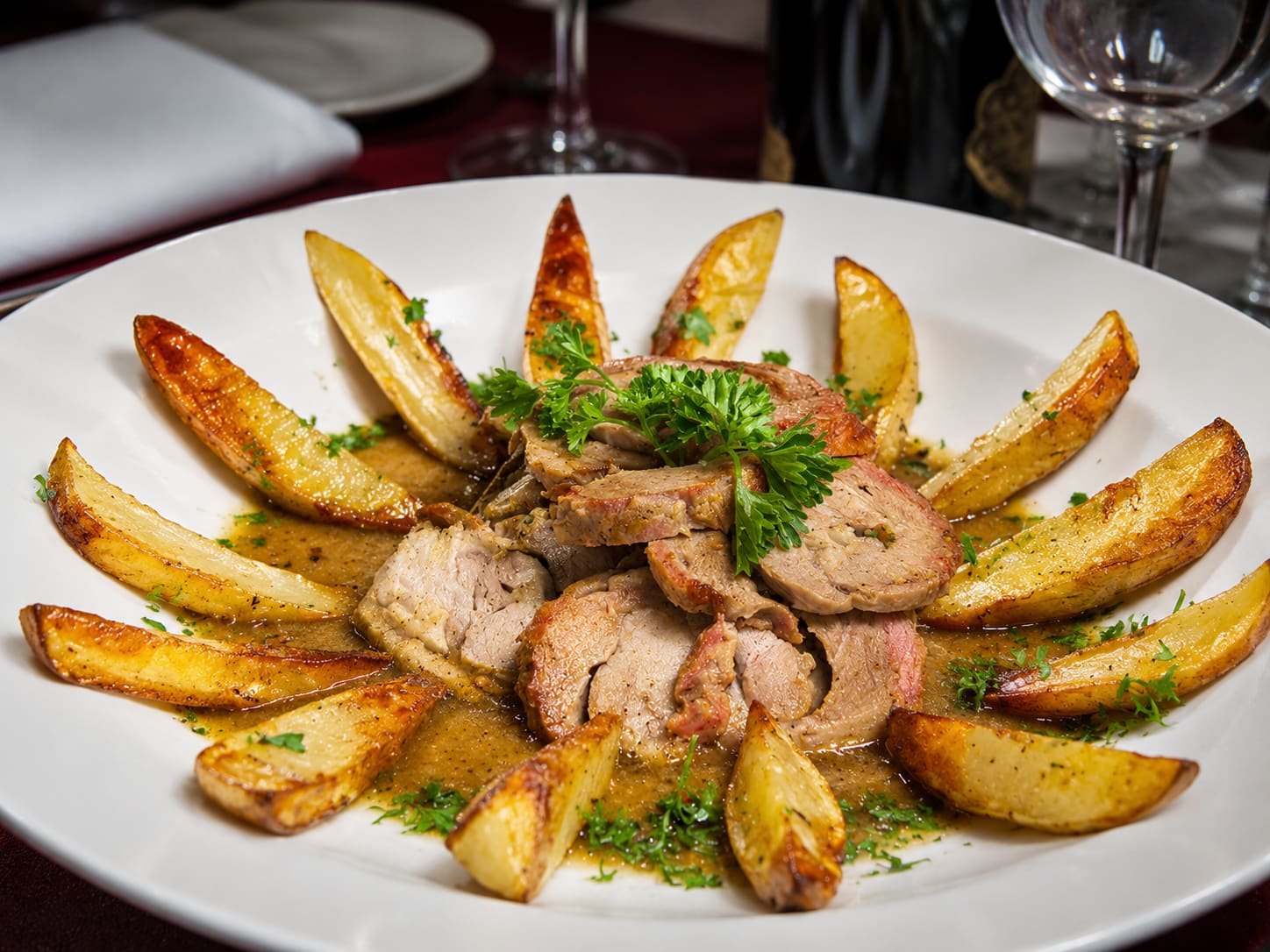 Veal braciolettini — rolled and sliced veal with prosciutto visible in each spiral, piled in pan juices and ringed by roasted potato wedges.